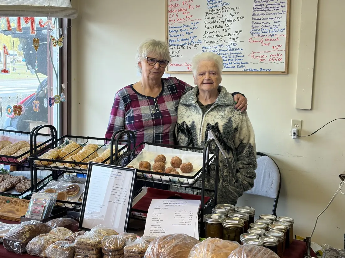 Two vendors standing behind a baked goods stall with breads, cookies and jars of preserves