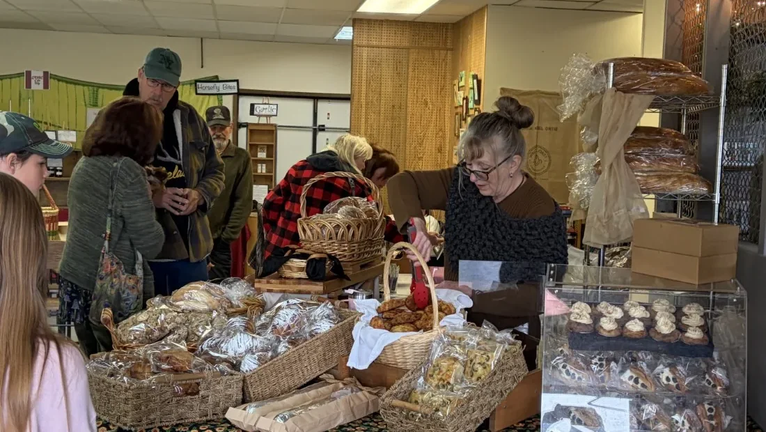 Vendors and customers at the market bakery counter, with baskets of bread and a display case of pastries