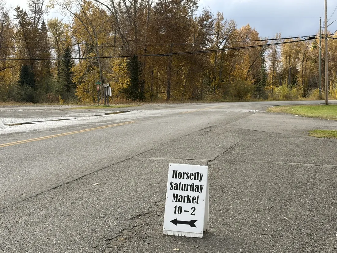 A-frame sign reading "Horsefly Saturday Market 10–2" with an arrow, set on the road beside autumn trees